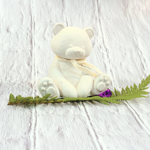 Small Afterstone teddy bear figurine sitting on a green fern leaf with a tiny purple flower, set against a white painted wood background.