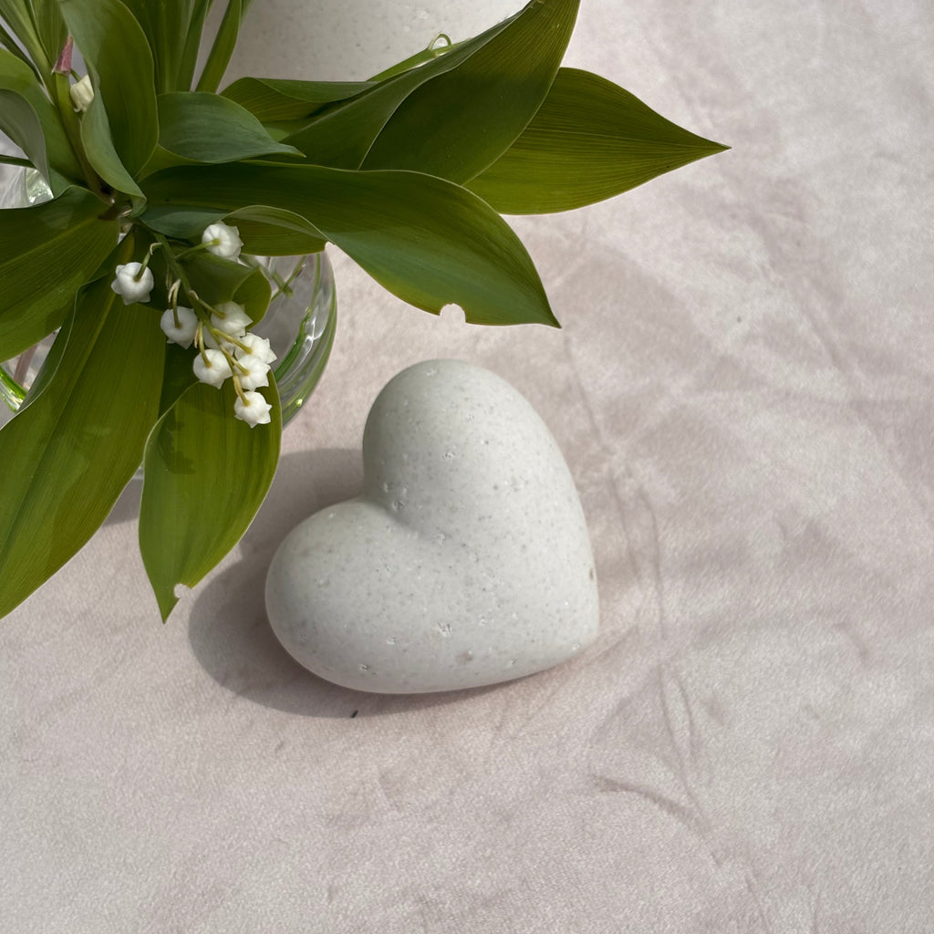 Heart-shaped stone on a textured surface with green leaves and small white flowers.