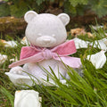 A small white Afterstone teddy bear figurine/ornament, made from ashes siting on green grass, wearing a pink velvet bow. White rose petals are scattered around it, with blurred green leaves and brown rocks in the background.