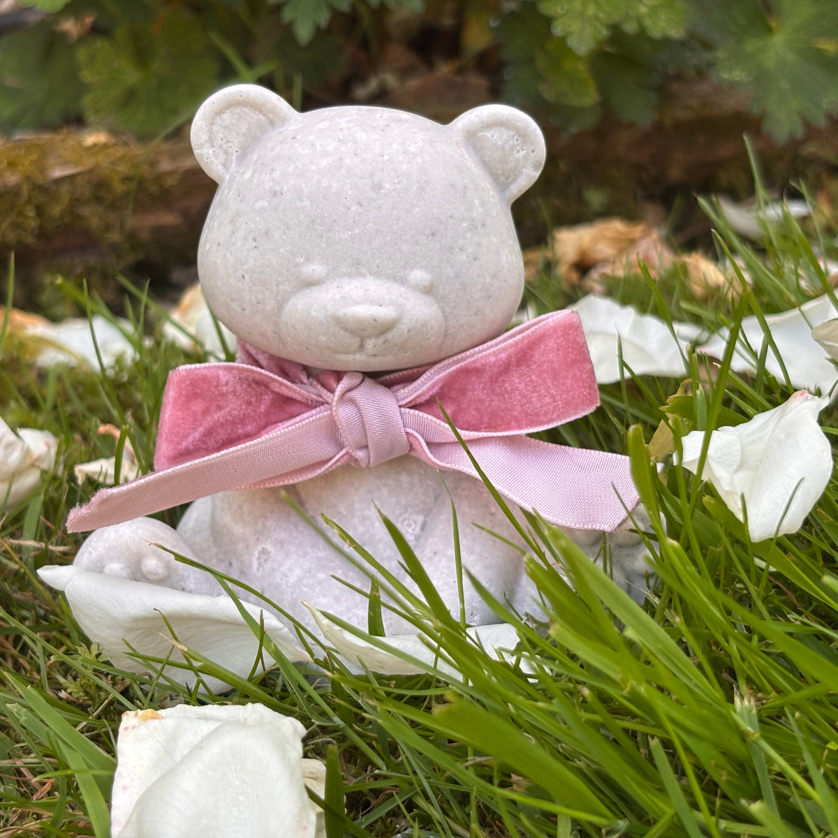 A small white Afterstone teddy bear figurine/ornament, made from ashes siting on green grass, wearing a pink velvet bow. White rose petals are scattered around it, with blurred green leaves and brown rocks in the background.
