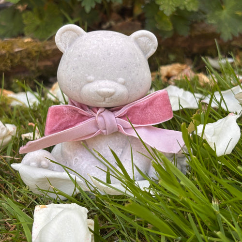 A small white Afterstone teddy bear figurine/ornament, made from ashes siting on green grass, wearing a pink velvet bow. White rose petals are scattered around it, with blurred green leaves and brown rocks in the background.