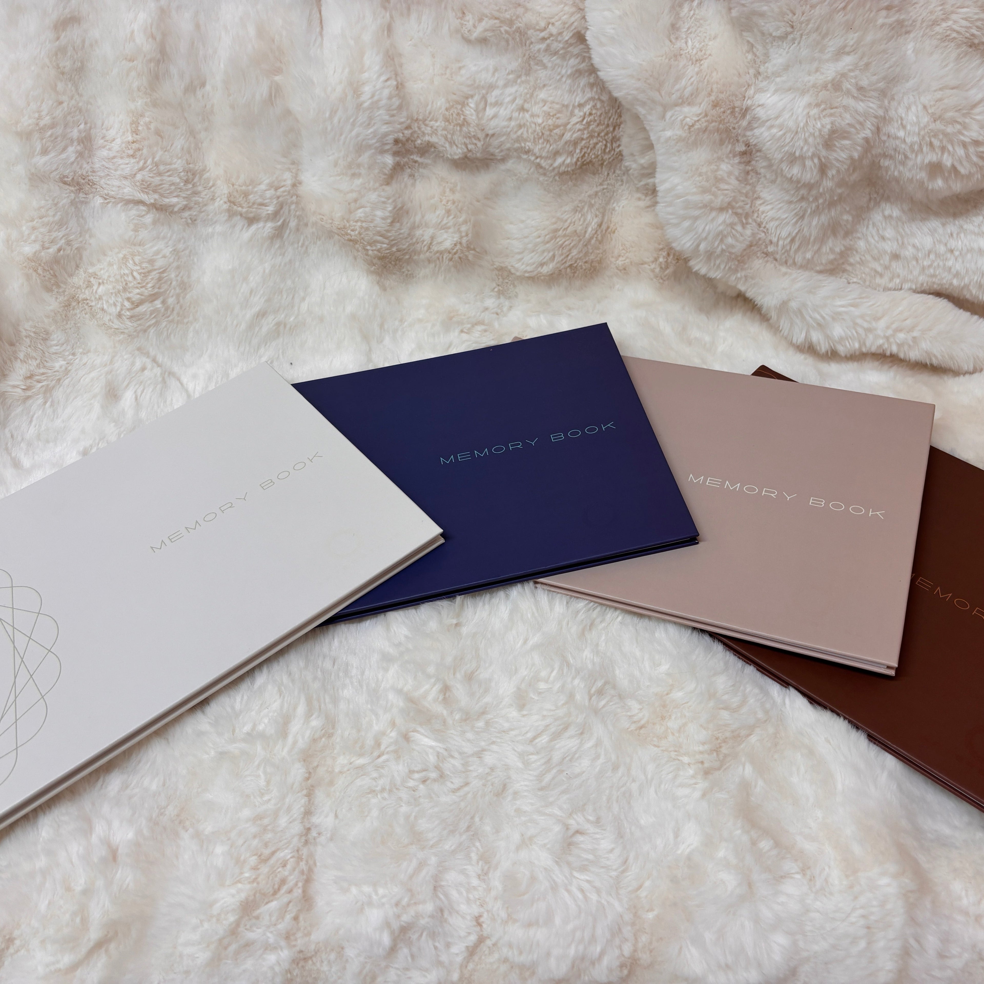 Four hardcover memory books in ivory/silve, navy blue/turquoise, beige/ivory, and chocolate/copper, arranged in a fan shape on a fluffy cream faux fur blanket. Each book features the text 'MEMORY BOOK' in a light-colored font with spot UV varnish.