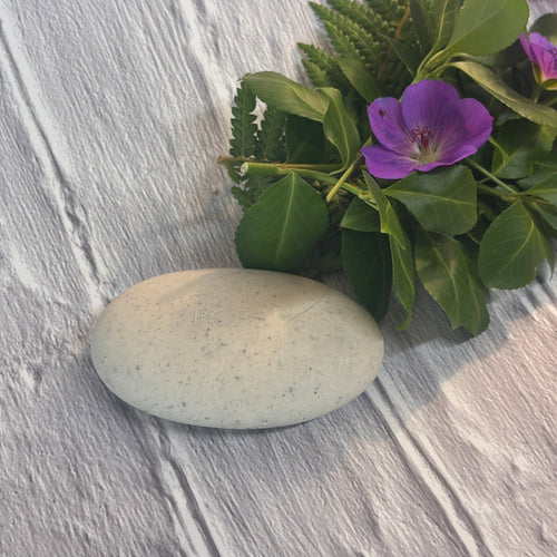 Afterstone solo pebble made from pet or human ashes on a brick-textured background with green foliage and purple flowers