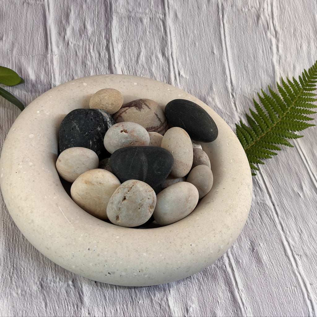 A round, shallow, cream-coloured ash into stone trinket bowl where you can see the ash flecks, filled with smooth actual river pebbles in neutral shades, styled with a green fern leaf on a white textured wooden background.