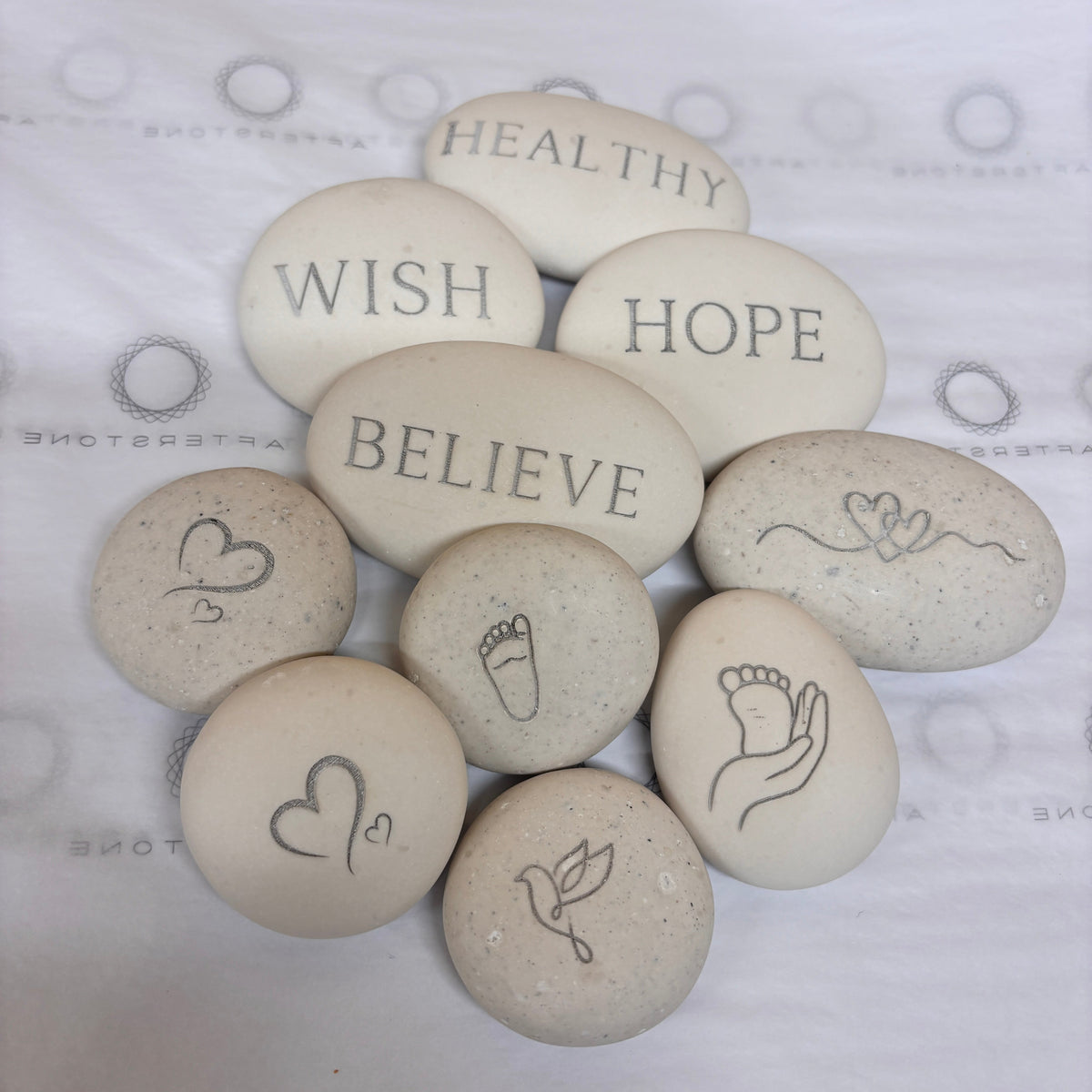 A group of round, light beige Afterstones made from pet or human ashes with engraved words and symbols/icons including 'HEALTHY', 'WISH', 'BELIEVE', 'HOPE', and icons including hand/foot icons, arranged on a white patterned fabric background.