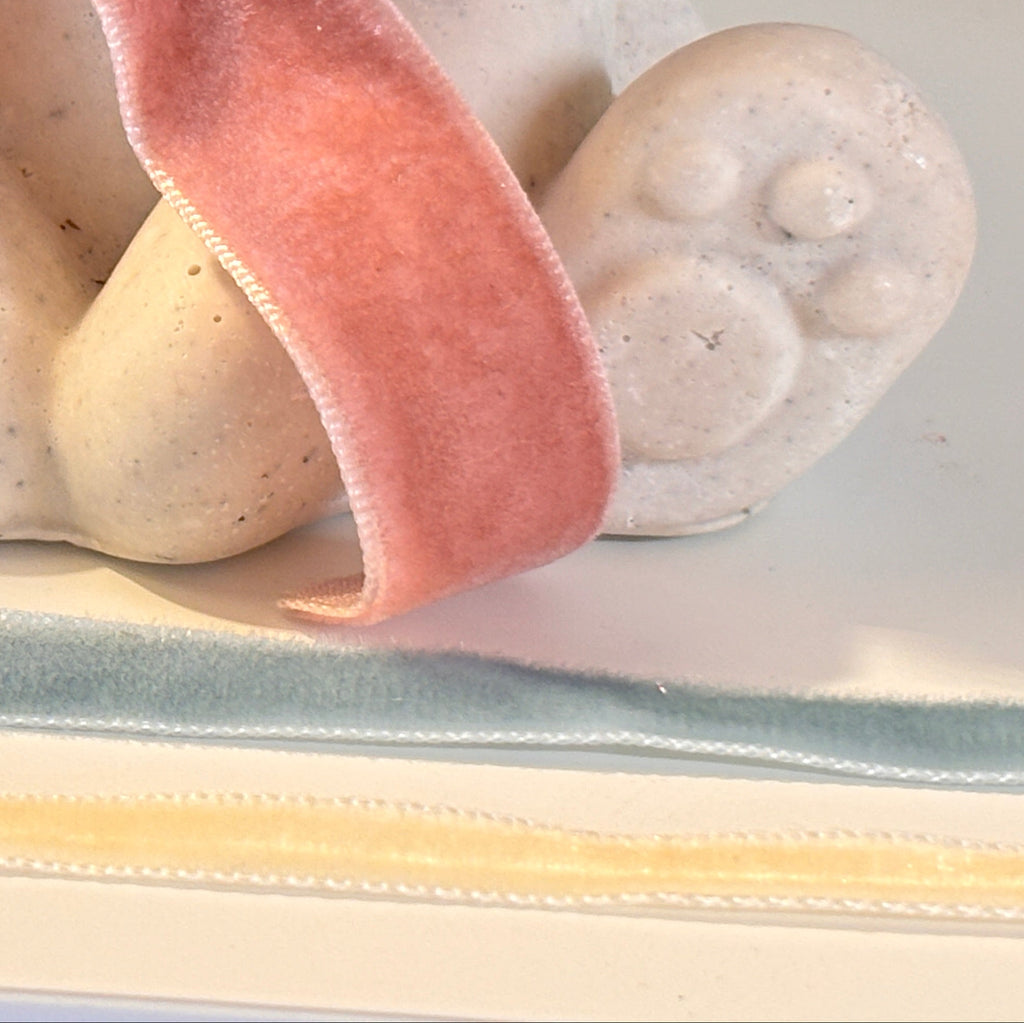 A close-up of a small white Afterstone teddy bear figurine with a soft pink velvet bow tied around its neck, sitting on a light surface with the other velvet ribbon alternatives showing is dusky blue and cream/oatmeal.