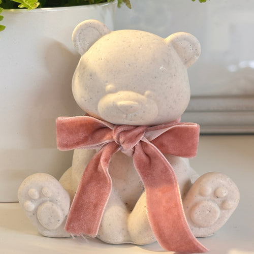 A small Afterstone teddy bear ornament, made from pet or human ashes with a speckled off-white stone-like finish, wearing a dusty pink velvet bow, displayed on a white surface with a potted fern in the background.