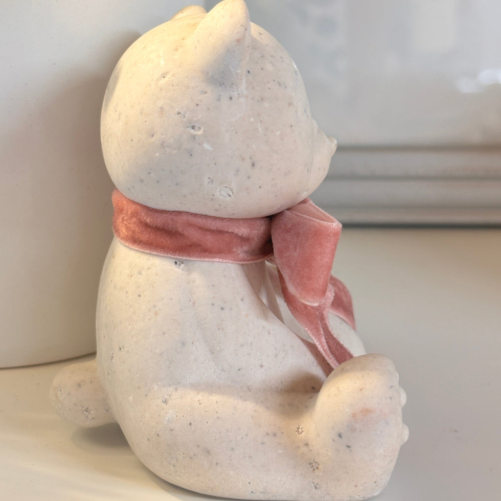 A small seated Afterstone teddy bear ornament/figurine in a speckled off-white stone material, wearing a soft pink scarf, photographed indoors on a light surface with a white planter in the background.