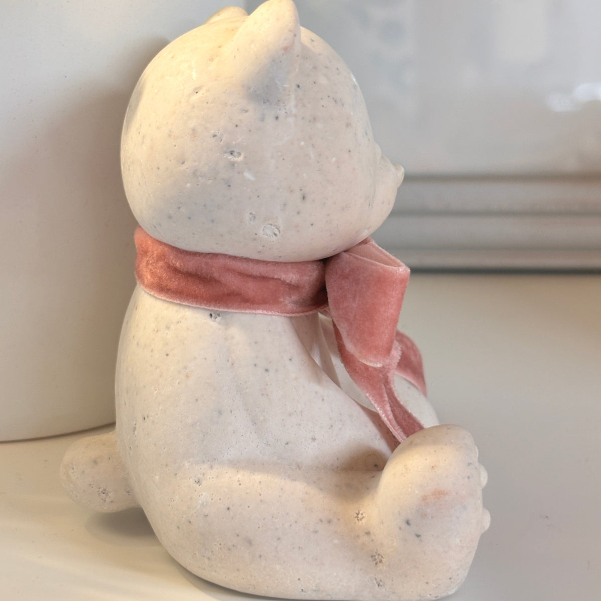 A small seated Afterstone teddy bear ornament/figurine in a speckled off-white stone material, wearing a soft pink scarf, photographed indoors on a light surface with a white planter in the background.