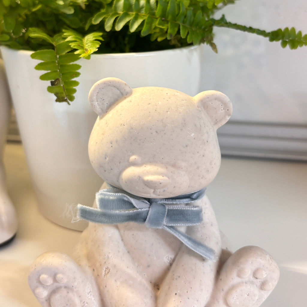 A small speckled cream-colored teddy bear figurine made from pet or human ashes, wearing a dusty blue ribbon bow, positioned in front of a white planter with a green fern.