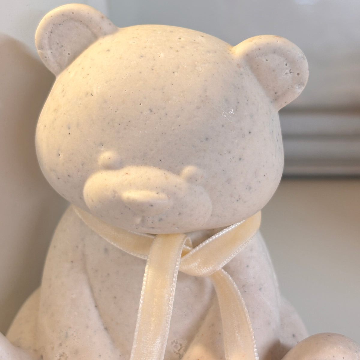 A close-up of an Afterstone speckled cream-colored teddy bear figurine , made with human or pet ashes, wearing a cream/oatmeal velvet ribbon scarf, set against a softly blurred background with a green fern in a pot.