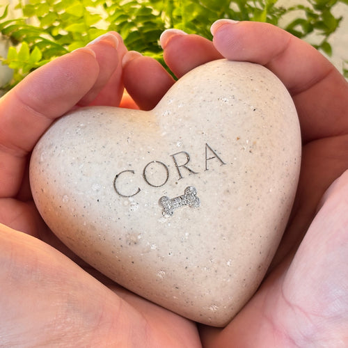 A personalised heart-shaped Afterstone memorial ornament, made from pet or human ashes engraved with the name 'CORA' and a small bone icon, held in two hands against a blurred green garden background.