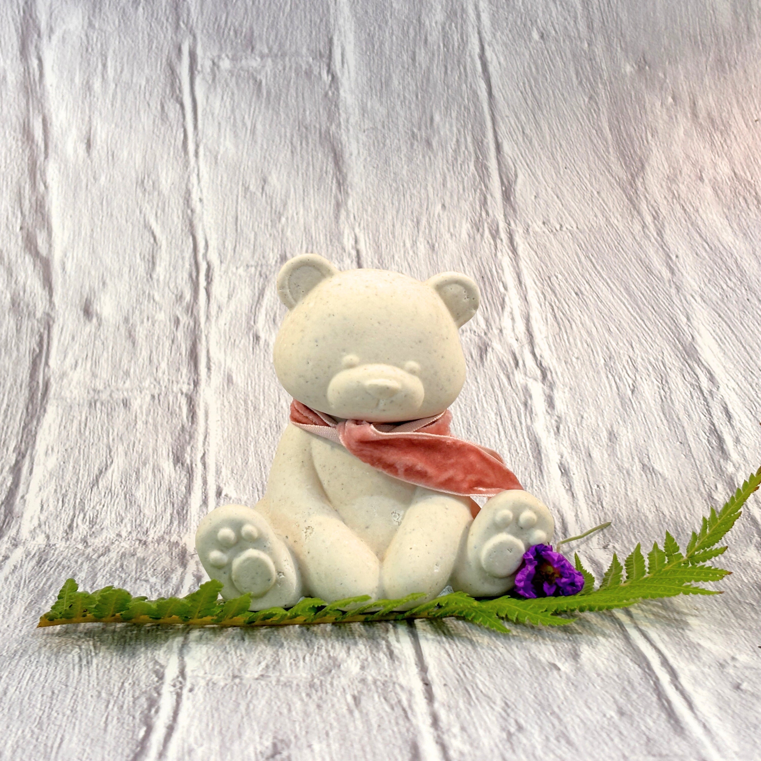 A small white Afterstone Ash-into-stone teddy bear figurine wearing a dusky pink scarf, sitting on a green fern leaf with a small purple flower, set against a white brick-textured background.