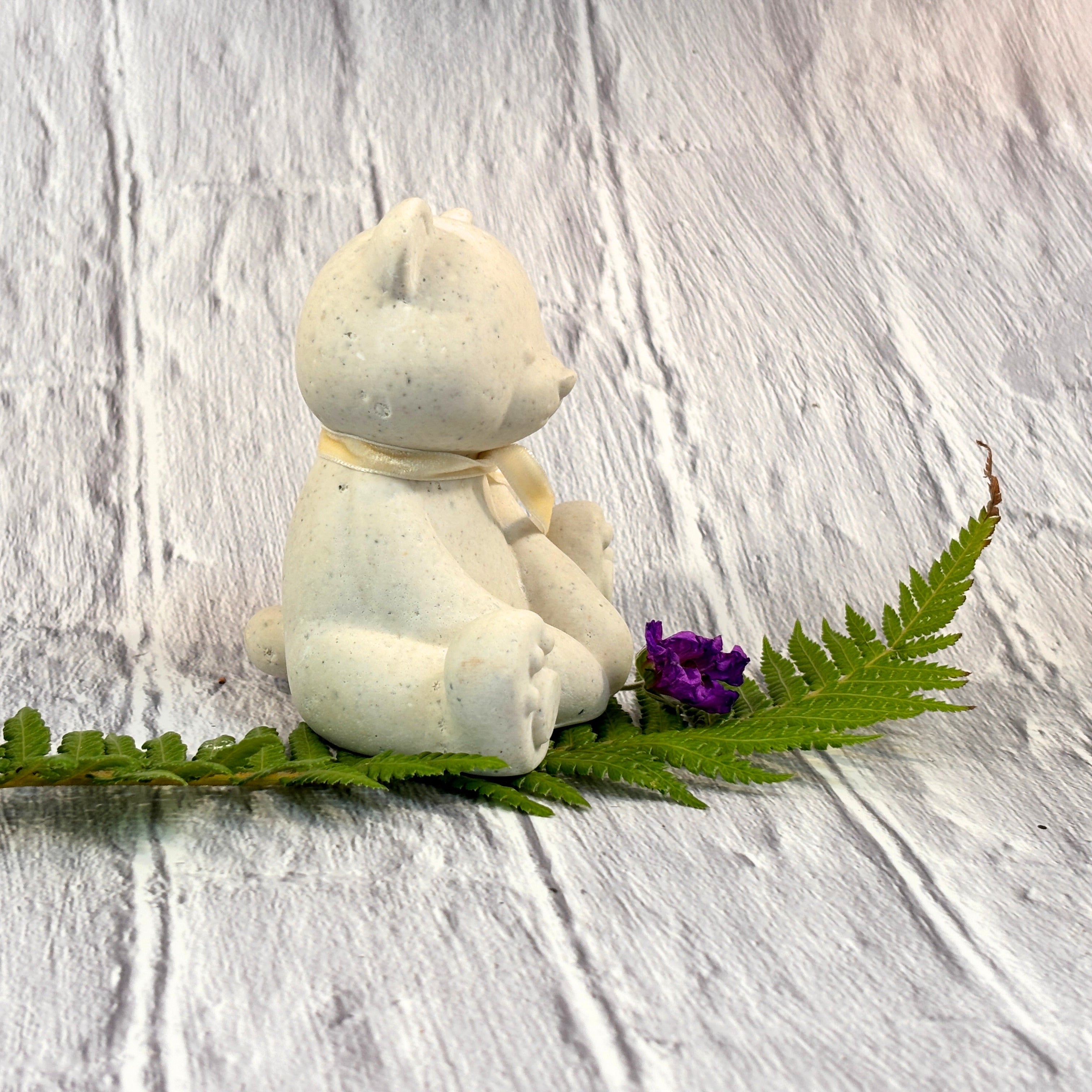 A small cream-coloured Afterstone (ash into-stone teddy bear figurine sitting on a green fern leaf next to a tiny purple flower, set against a white textured wooden background.