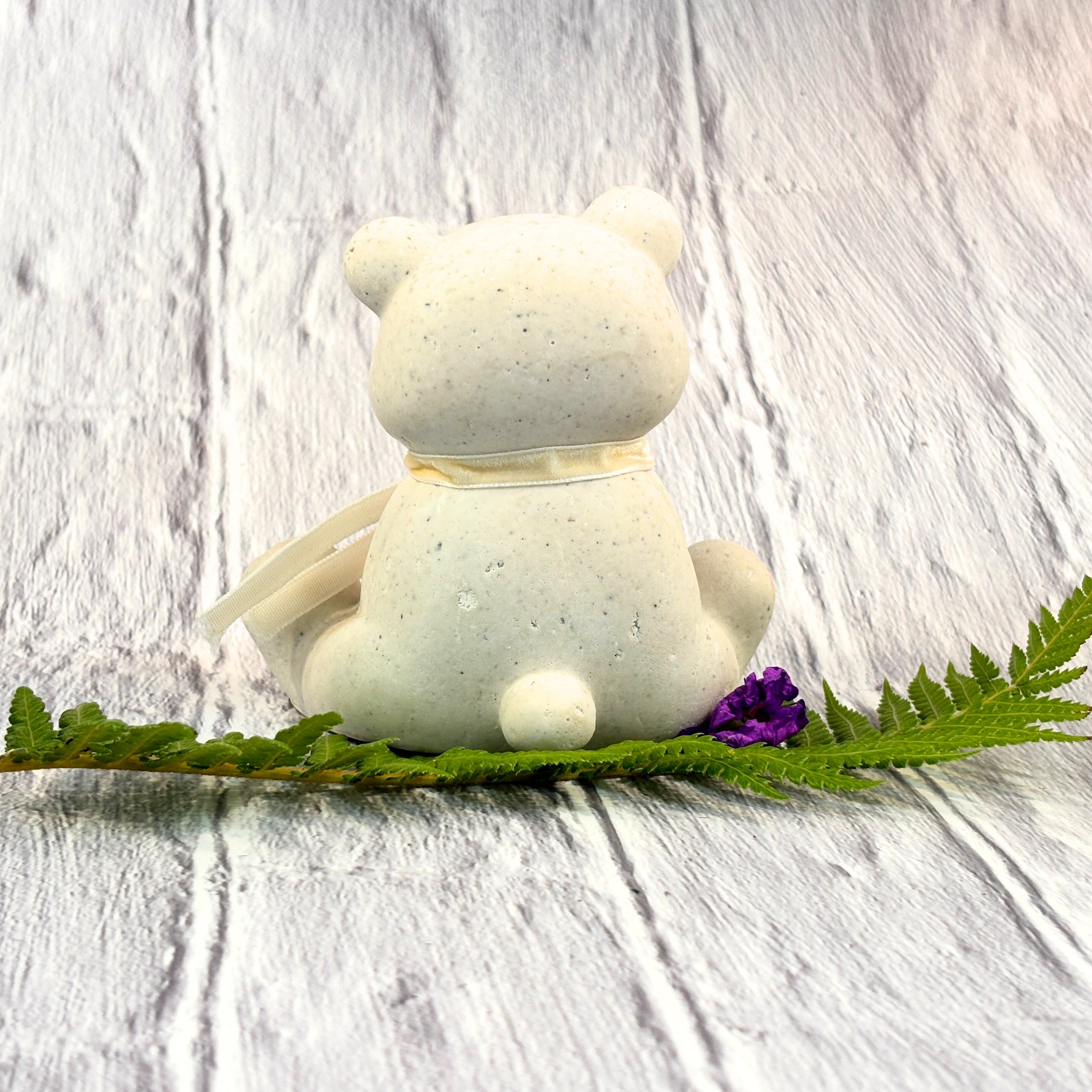 Cream Afterstone (ash-into-stone) teddy bear figurine with a velvet ribbon scarf, sitting, taken from behind. He is sitting on a fern branch with a purple flower 